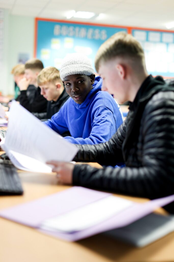 Functional Skills maths - Students work at computers in a business classroom, one holding a sheet of paper, Stockton Sixth Form College.