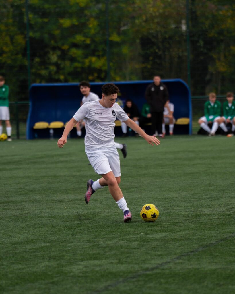 Sport (Level 2) - A footballer in white kit running with the ball past a dugout where substitutes are seated.