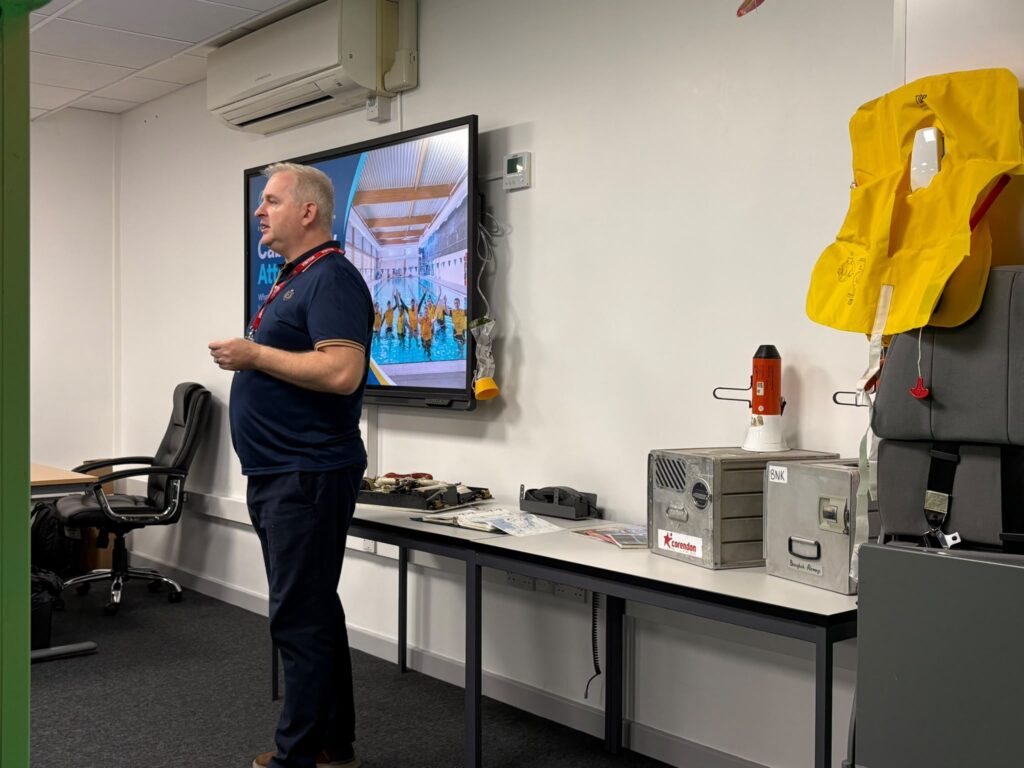 A trainer talks to the class, with safety equipment and a life jacket beside him.